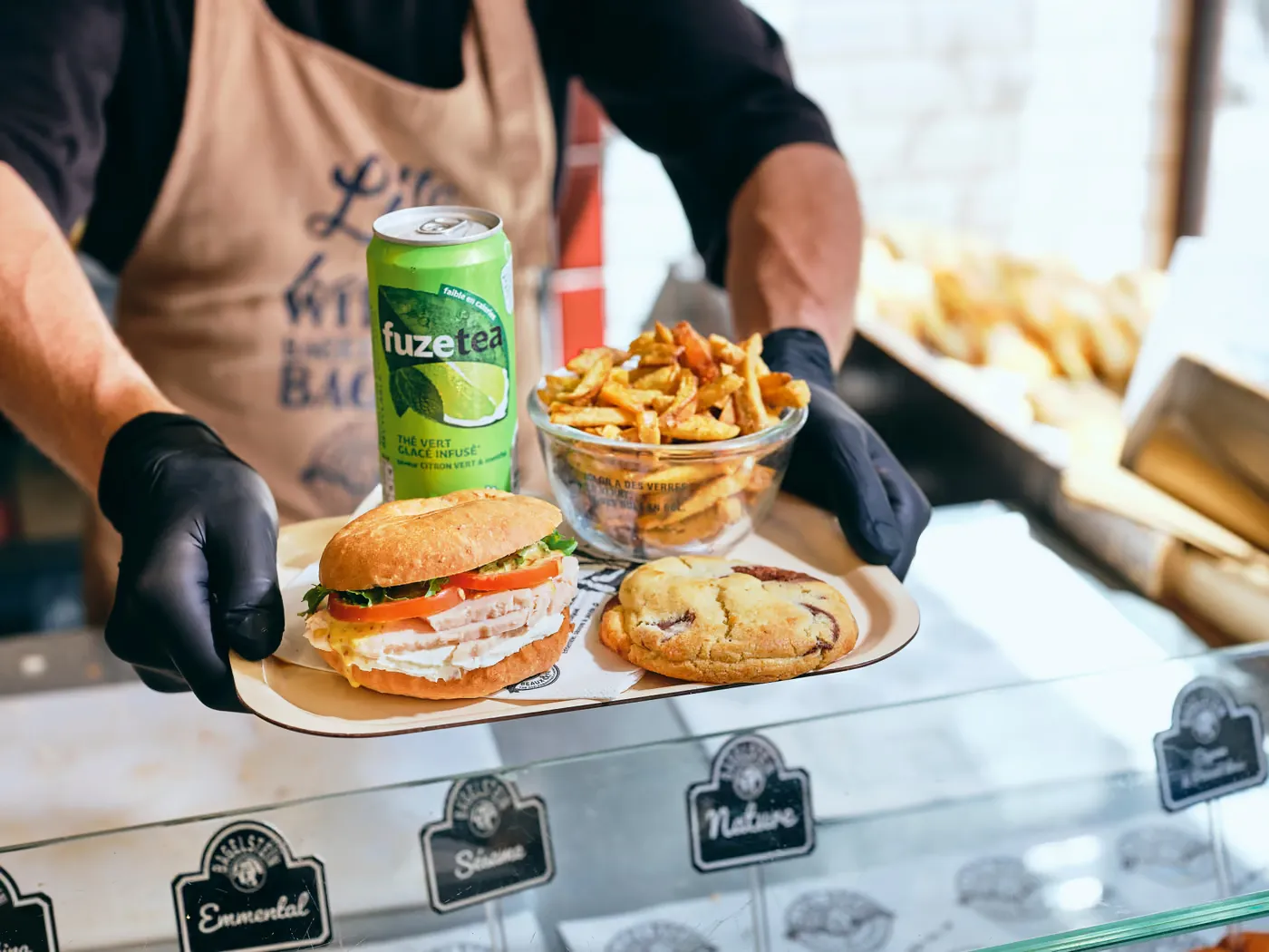 Un équipier présente un plateau (bagel, frites, cookie et boisson) par-dessus la vitrine